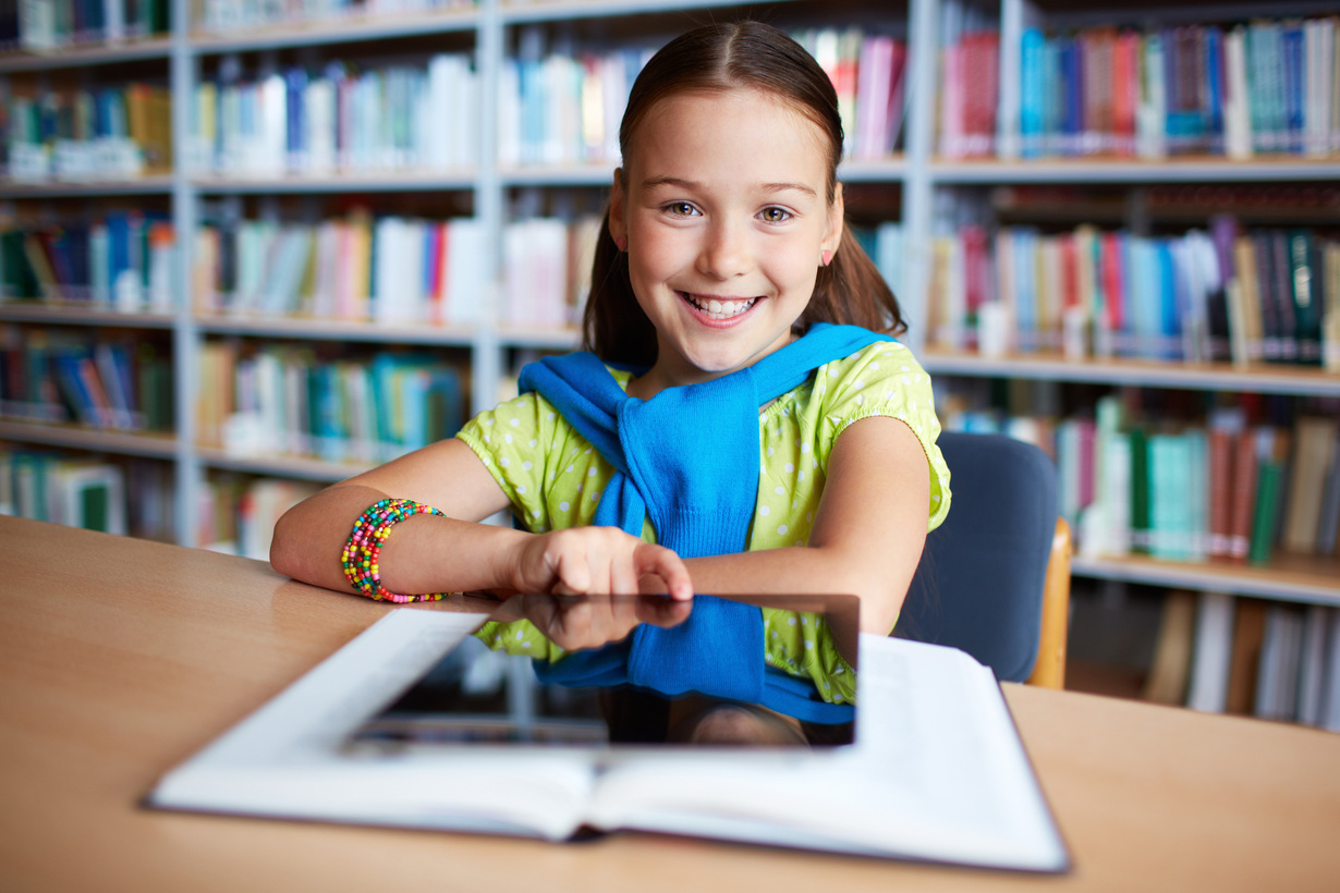 Hintergrundbild: Lächelndes Mädchen mit einem Tablet vor sich an einer Schulbank sitzend vor Bücherregal im Hintergrund
