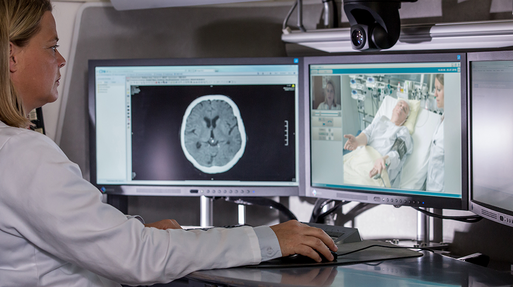 Medizinerin betrachtet Brain-Scan und überwacht Patienten-Video auf Monitoren.