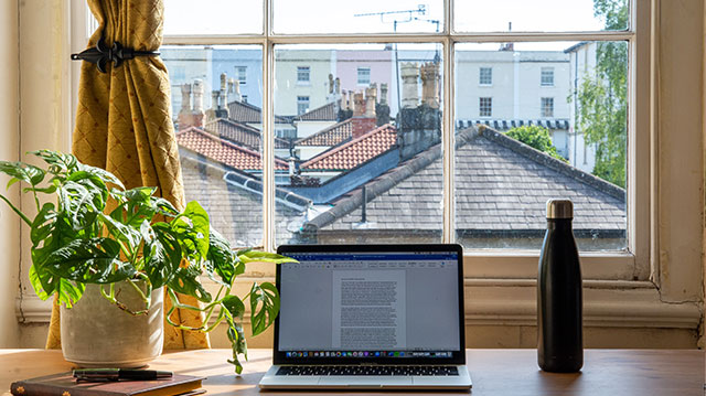 Schreibtisch mit Rechner, Pflanze, Notizbuch und Trinkflasche vor Fenster mit Aussicht auf Häuserdächer und einen Wohnblock